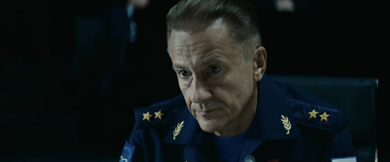 Invasion (2020) movie still: closeup — A dimly lit room with a man in a formal military uniform sitting down. The man keeps his head bowed while…