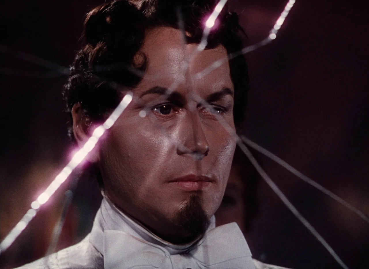 The Tales of Hoffmann (1951) movie still: closeup — A man in formal wear stands behind a glass surface that has shattered radially. Sharp lines of…