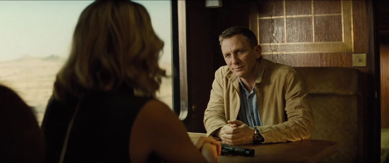 Spectre (2015) movie still: medium — An interior train compartment with dark wood paneling and a side window view of desert. A man in a tan jacket…