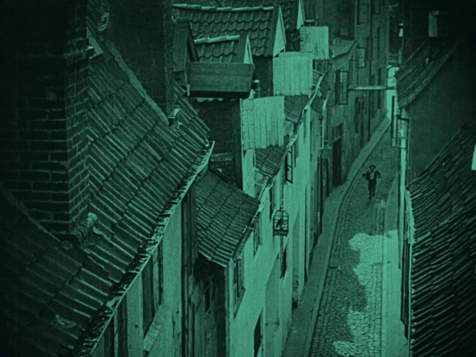 Nosferatu (1922) movie still: wide — A steep-angled view looks down into a narrow, dimly lit cobblestone alleyway. A lone figure in dark clothes…