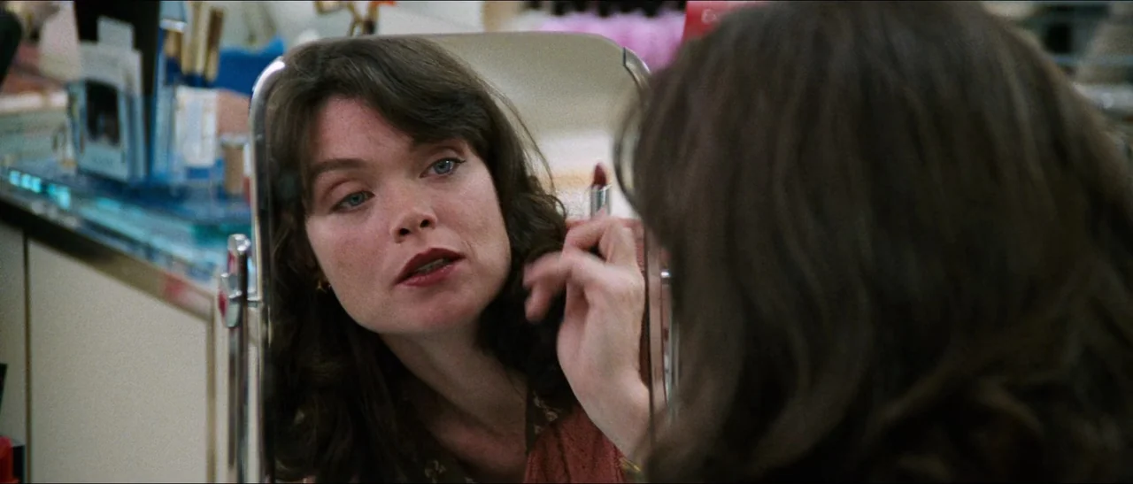 Election (1999) movie still: closeup — A woman stands before a vanity mirror in a busy retail environment. She holds a tube of lipstick and applies…