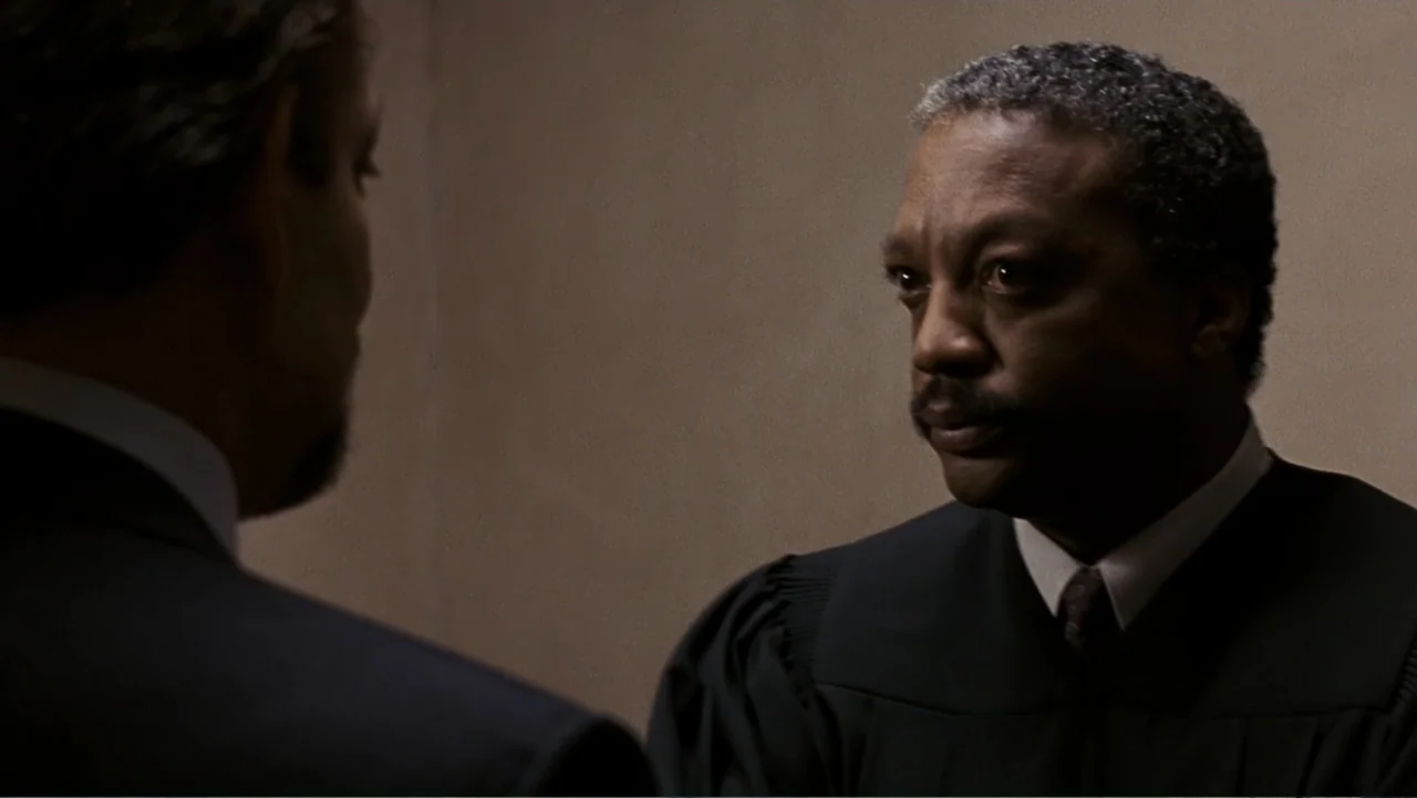 Presumed Innocent (1990) movie still: medium — An interior space with plain beige walls and muted lighting. A judge in robes speaks to a man in a…