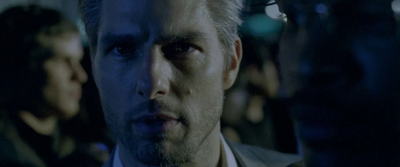 Collateral (2004) movie still: closeup — A dark, crowded space is dimly lit by blue ambient light. A man stares intently forward while slightly…