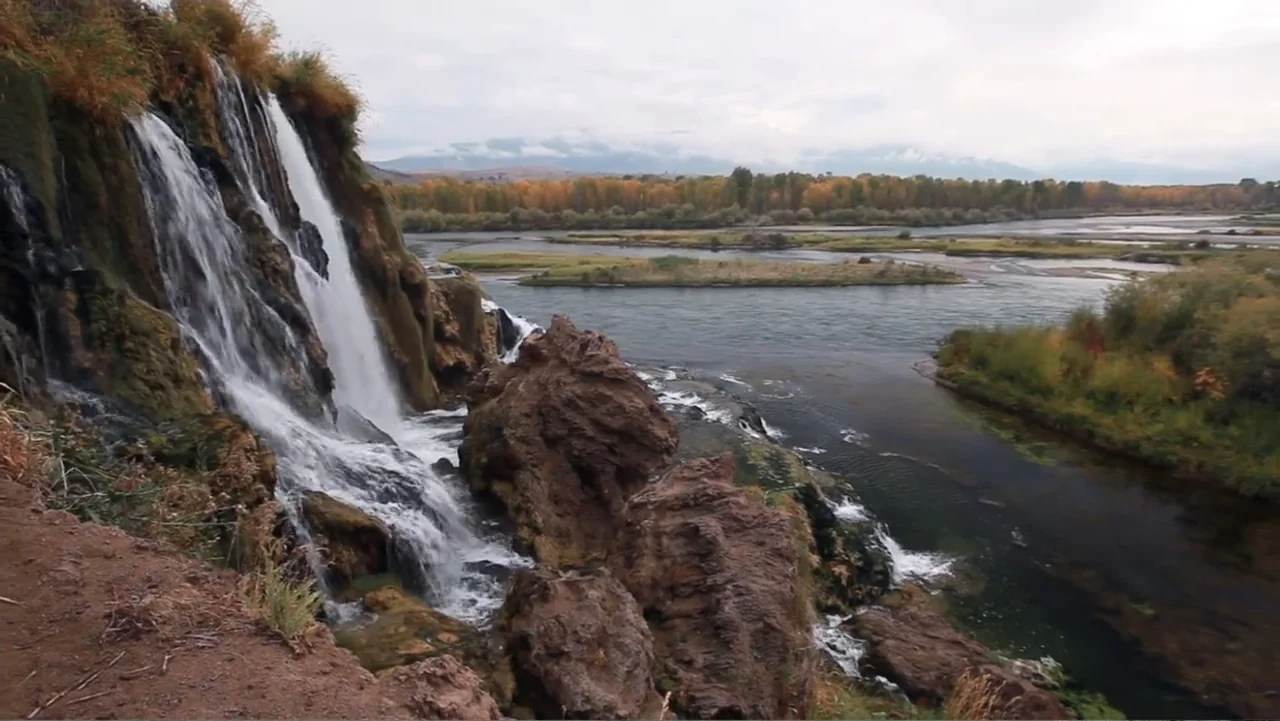Idaho: The Movie (2012) movie still: wide — A rugged cliff face with a cascading waterfall overlooks a wide, winding river. The surrounding…