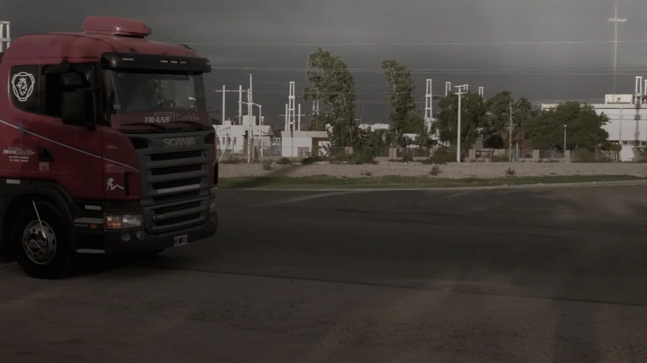Still Life (2013) movie still: medium — A red semi-truck sits on the paved shoulder of a road near industrial power lines. The truck is stationary…