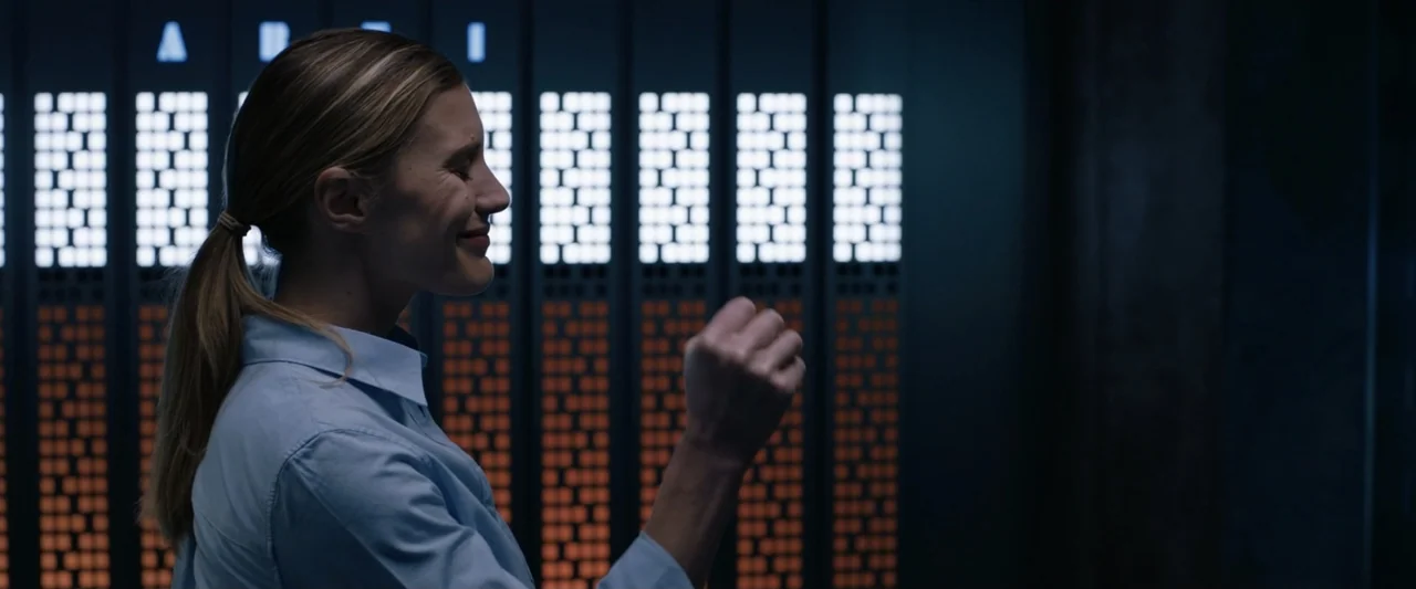 2036 Origin Unknown (2018) movie still: medium — A woman in a blue shirt stands before a wall of glowing server racks. She maintains a side profile…