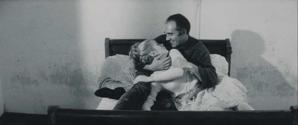 Les Créatures (1966) movie still: medium — A sparsely furnished room contains a simple wooden bed frame with light bedding. A man embraces a woman…