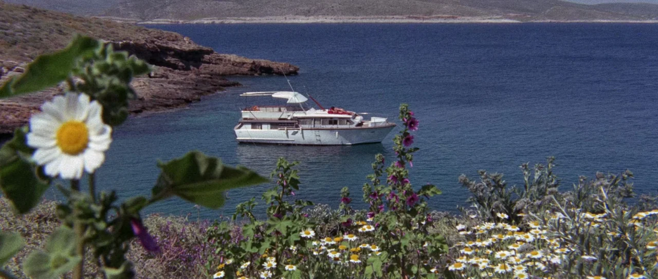 The Case of the Scorpion’s Tail (1971) movie still: wide — A white motor yacht floats in a calm blue bay near a rocky coastline. Wildflowers bloom…