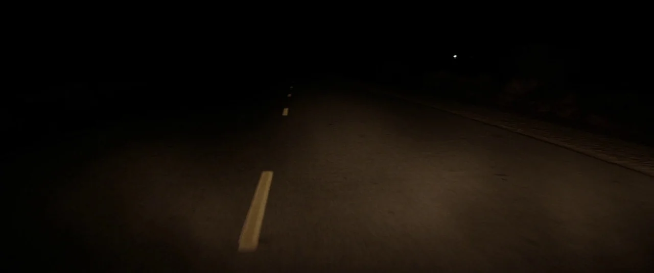 Southbound (2015) movie still: establishing — A dark asphalt road extends into the night with minimal visibility. Faded white lane markings run…