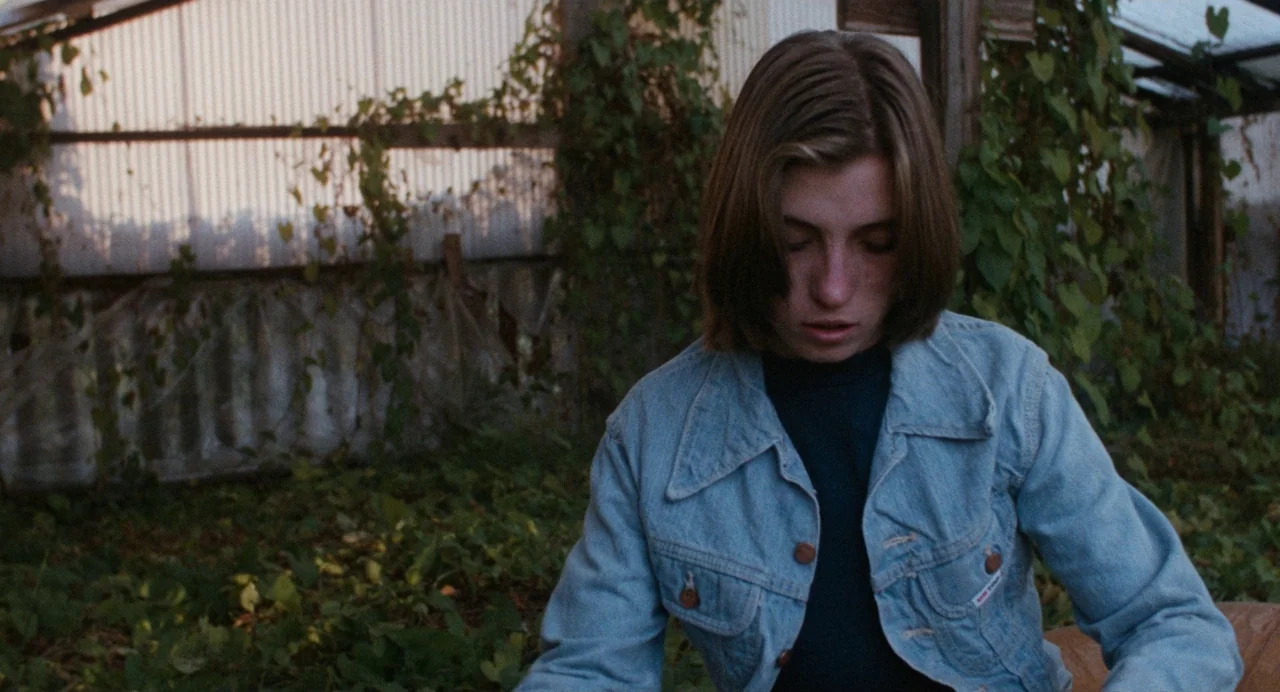Out of the Blue (1980) movie still: medium — A neglected greenhouse contains overgrown plants with a corrugated metal rear wall. A young woman looks…