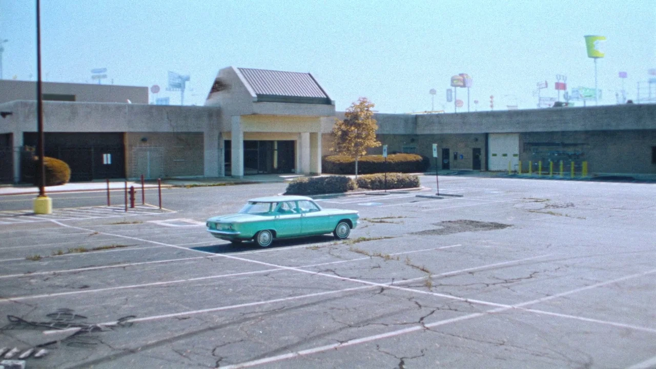 Strawberry Mansion (2021) movie still: wide — A vast, empty concrete parking lot sits before a low-slung, beige commercial building facade. A single…