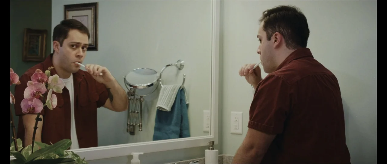 Ordinary Things (2001) movie still: medium — A residential bathroom with pale walls and a large vanity mirror reflecting the subject. A man stands…