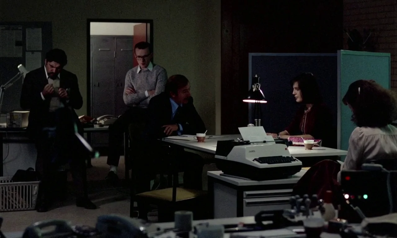 The Lost Honour of Katharina Blum (1975) movie still: wide — An open plan office space at night features desks, metal cabinets, and fluorescent…