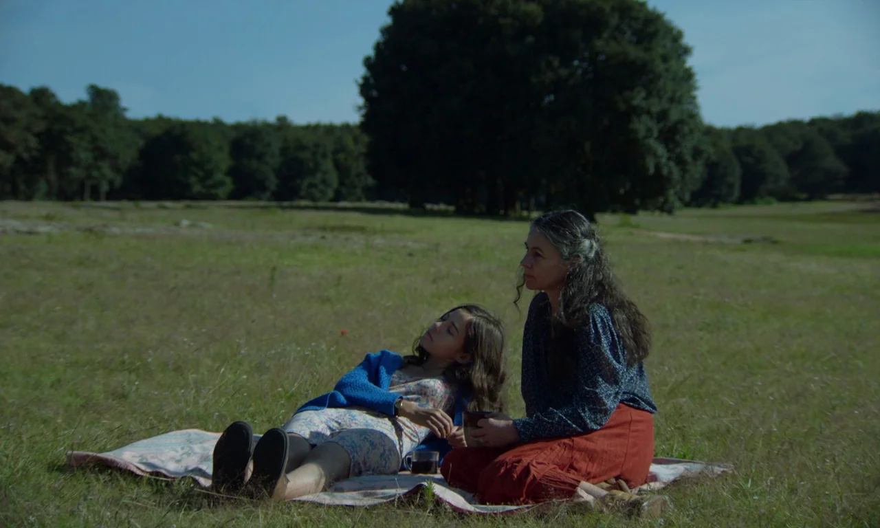 The Untamed (2016) movie still: wide — A woman and a girl rest on a quilt spread across a wide grassy field. Large trees line the horizon under a…