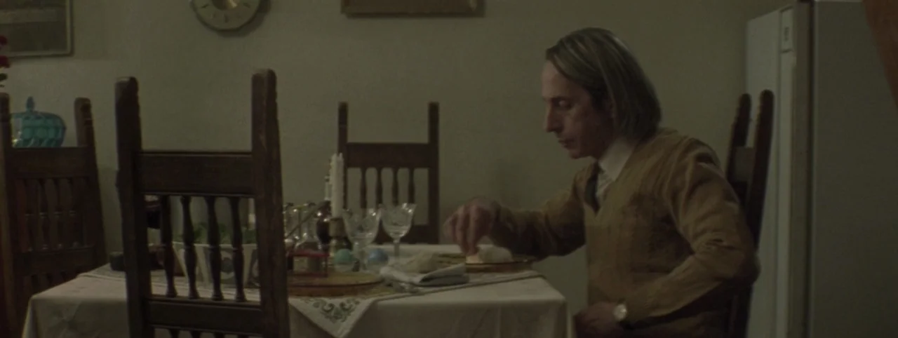 Post Mortem (2010) movie still: medium — A dimly lit dining room features wood furniture and a white patterned tablecloth. A man sits alone at the…