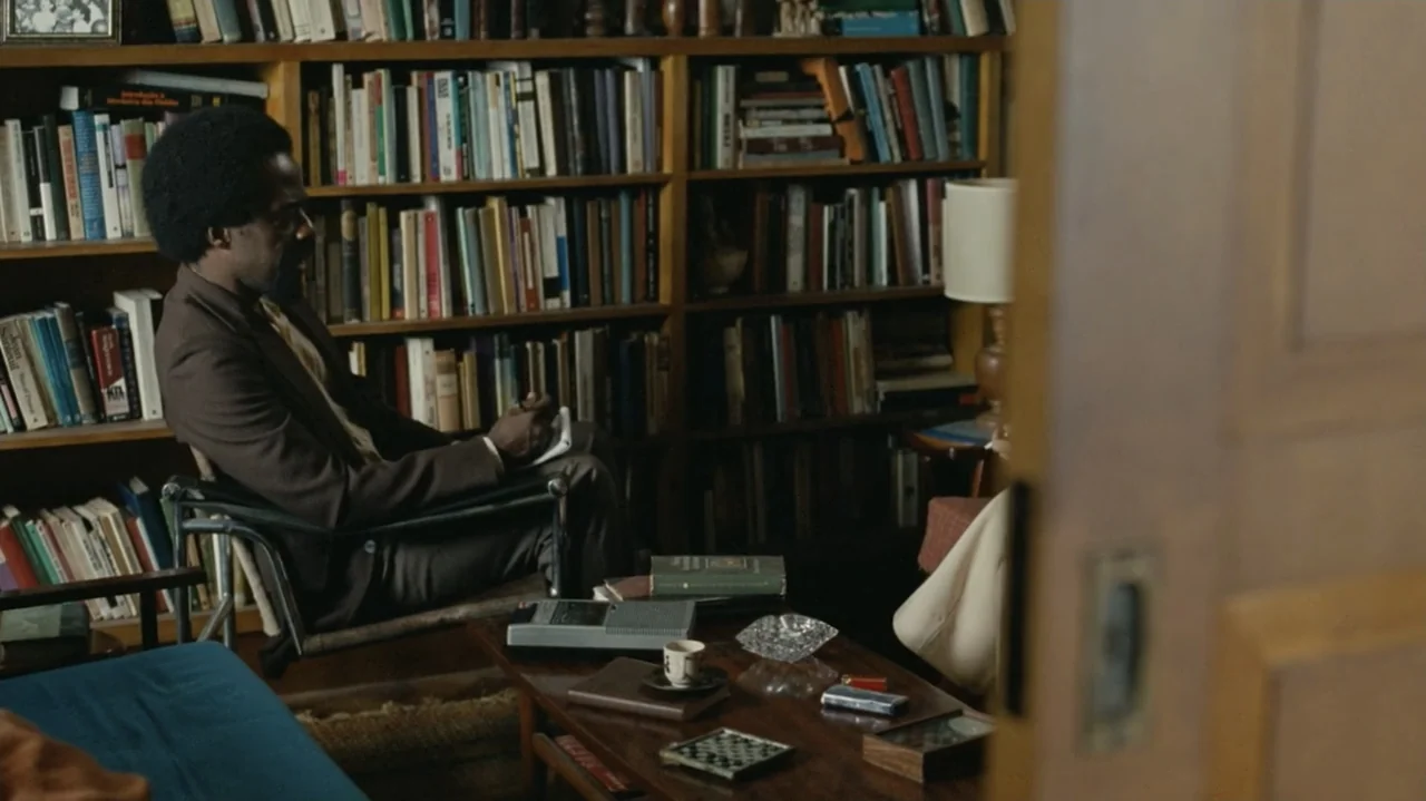 I'm Still Here (2024) movie still: medium — A study filled with overflowing bookshelves and a wooden table holds small personal items. A man sits in…