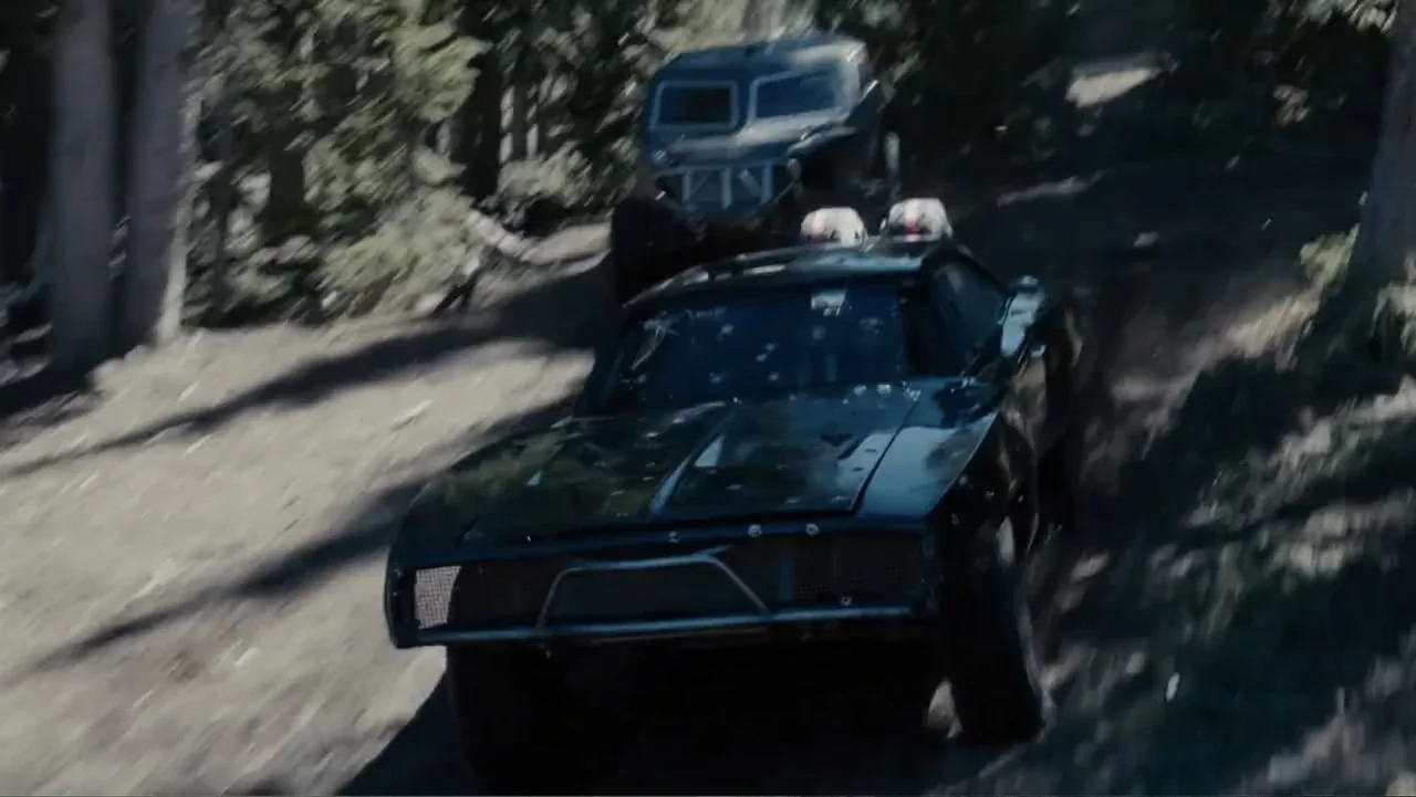 Furious 7 (2015) movie still: wide — A dark muscle car follows a heavily modified off-road vehicle along a dusty trail. Tall pine trees line the…