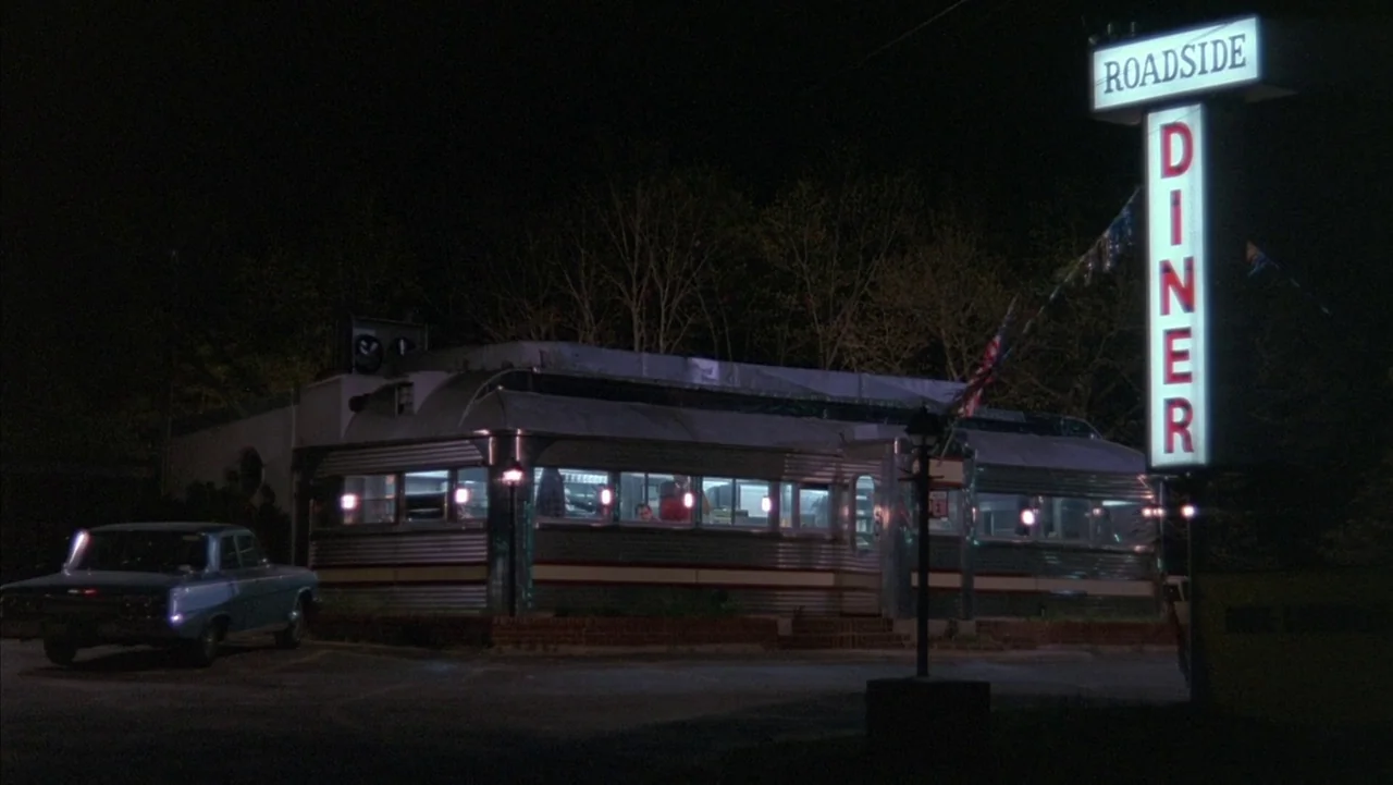 Baby It’s You (1983) movie still: establishing — A metal-clad roadside diner sits under a dark night sky surrounded by bare trees. An illuminated…