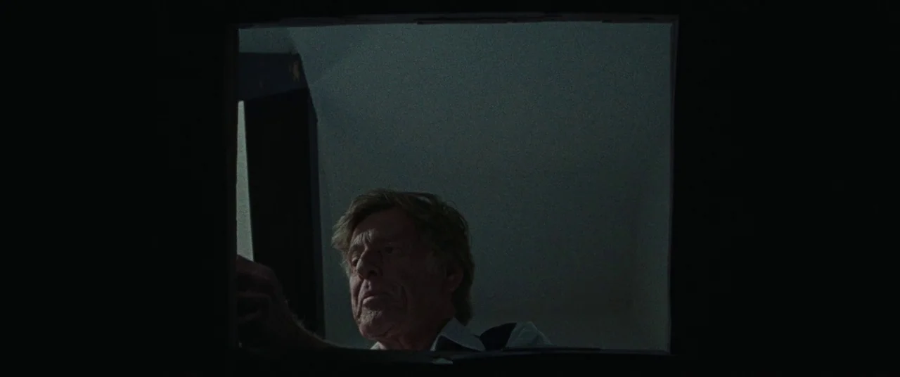 The Old Man & The Gun (2018) movie still: closeup — An older man peers upward through a dark, square cutout in a dim attic space. He stretches his…