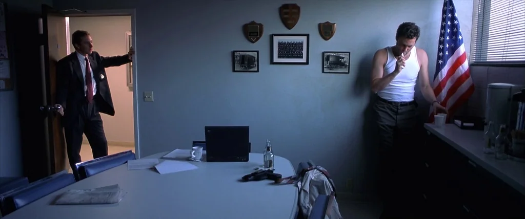 The Cell (2000) movie still: wide — A dimly lit room with grey walls features a central conference table and wall hangings. One man stands by an…