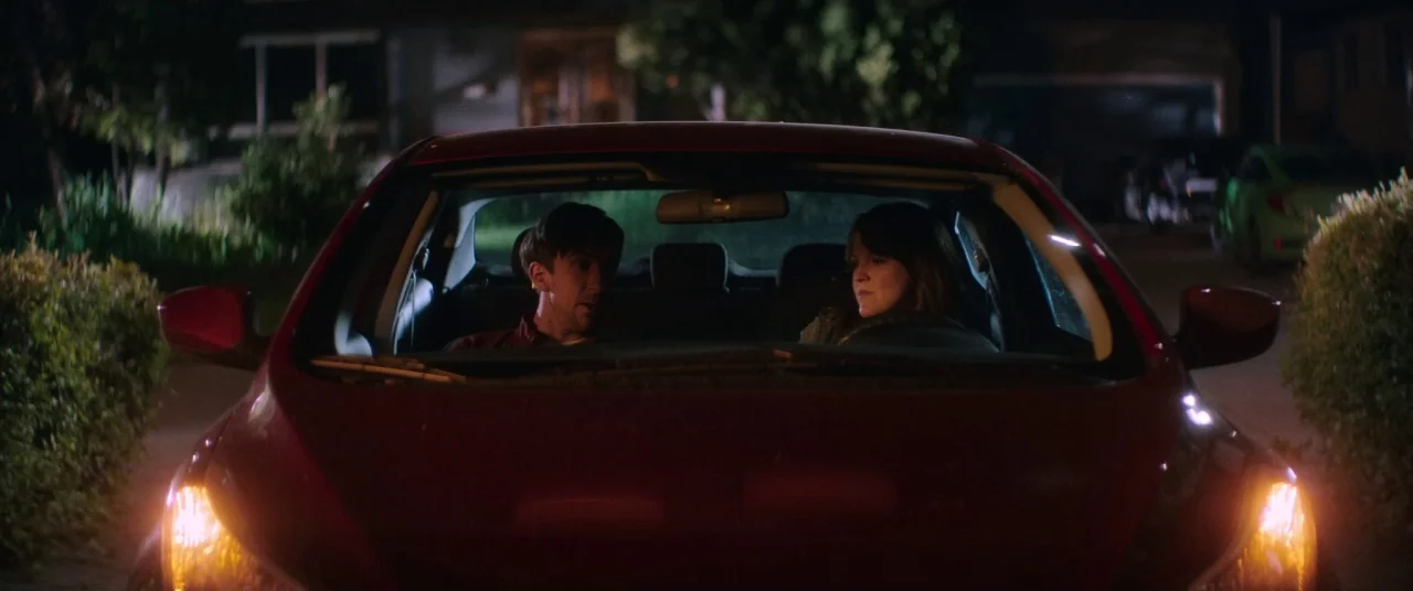 Room For Rent (2017) movie still: medium — A dark, quiet suburban street at night frames a red sedan parked with headlights on. A man and a woman…
