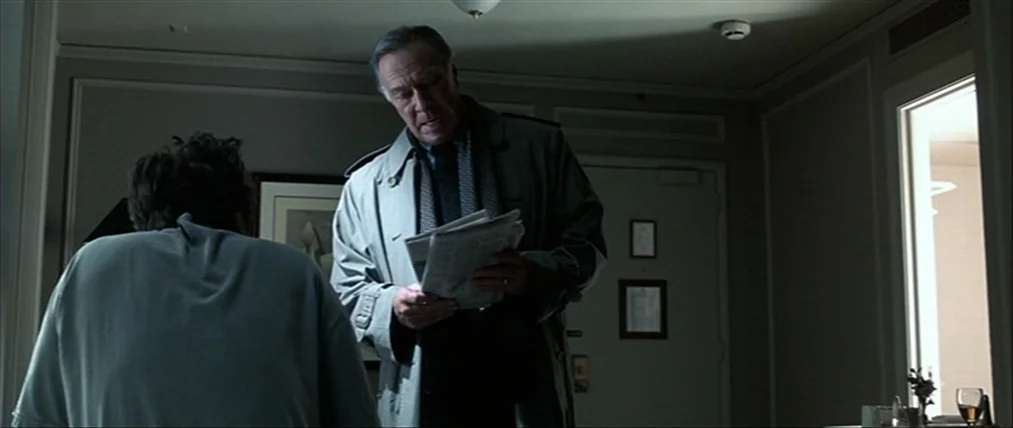 The Insider (1999) movie still: medium — A dimly lit hotel room with neutral walls and a doorway visible in background. An older man in a trench…