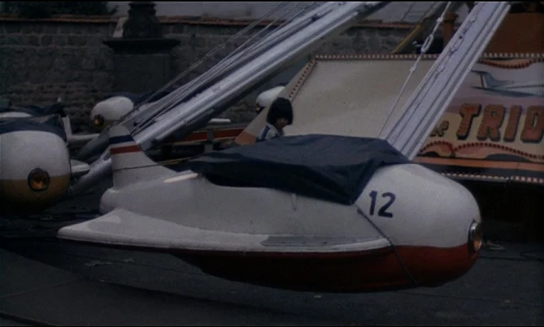 Small Change (1976) movie still: medium — An outdoor amusement park features a white airplane ride labeled with the number twelve. A child sits…
