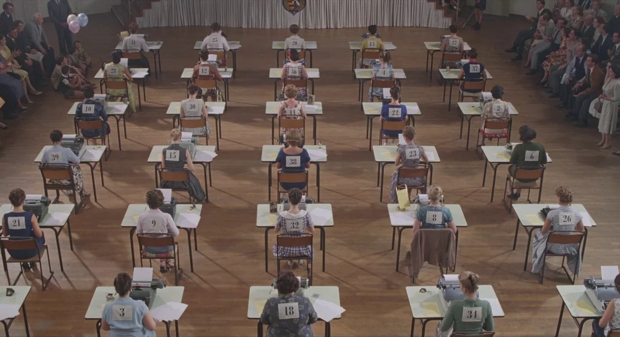 Populaire (2012) movie still: wide — A large hall contains rows of women seated at desks with numbered labels. Each woman operates a vintage…