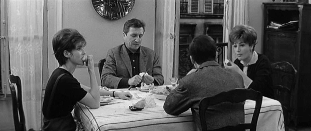 Antoine and Colette (1962) movie still: medium — A 1960s dining room features a set table with a white cloth and wooden furniture. Two men and two…