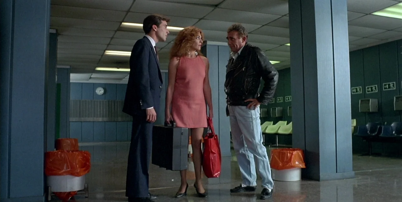 Law of Desire (1987) movie still: fullbody — A spacious, sterile room features blue columns and rows of plastic seating against walls. Three people…
