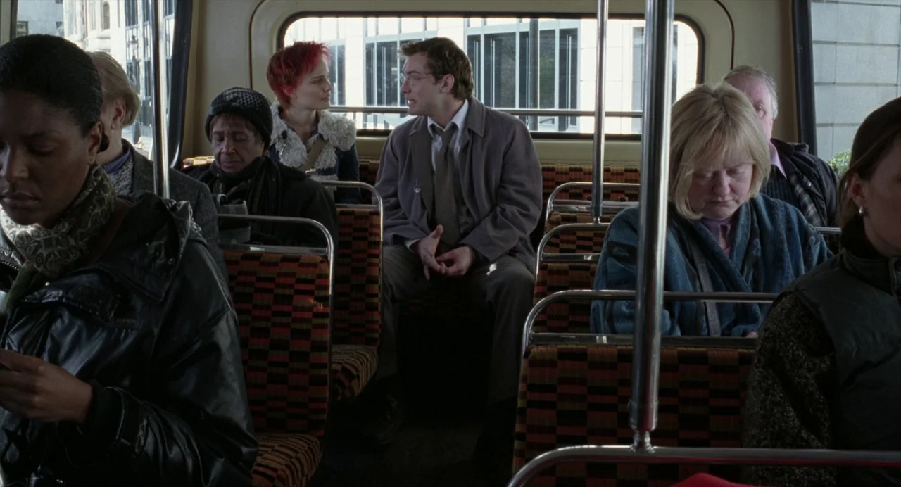 Closer (2004) movie still: wide — The interior of a public transit bus features checkered fabric seats and metal poles. A man and a woman sit facing…