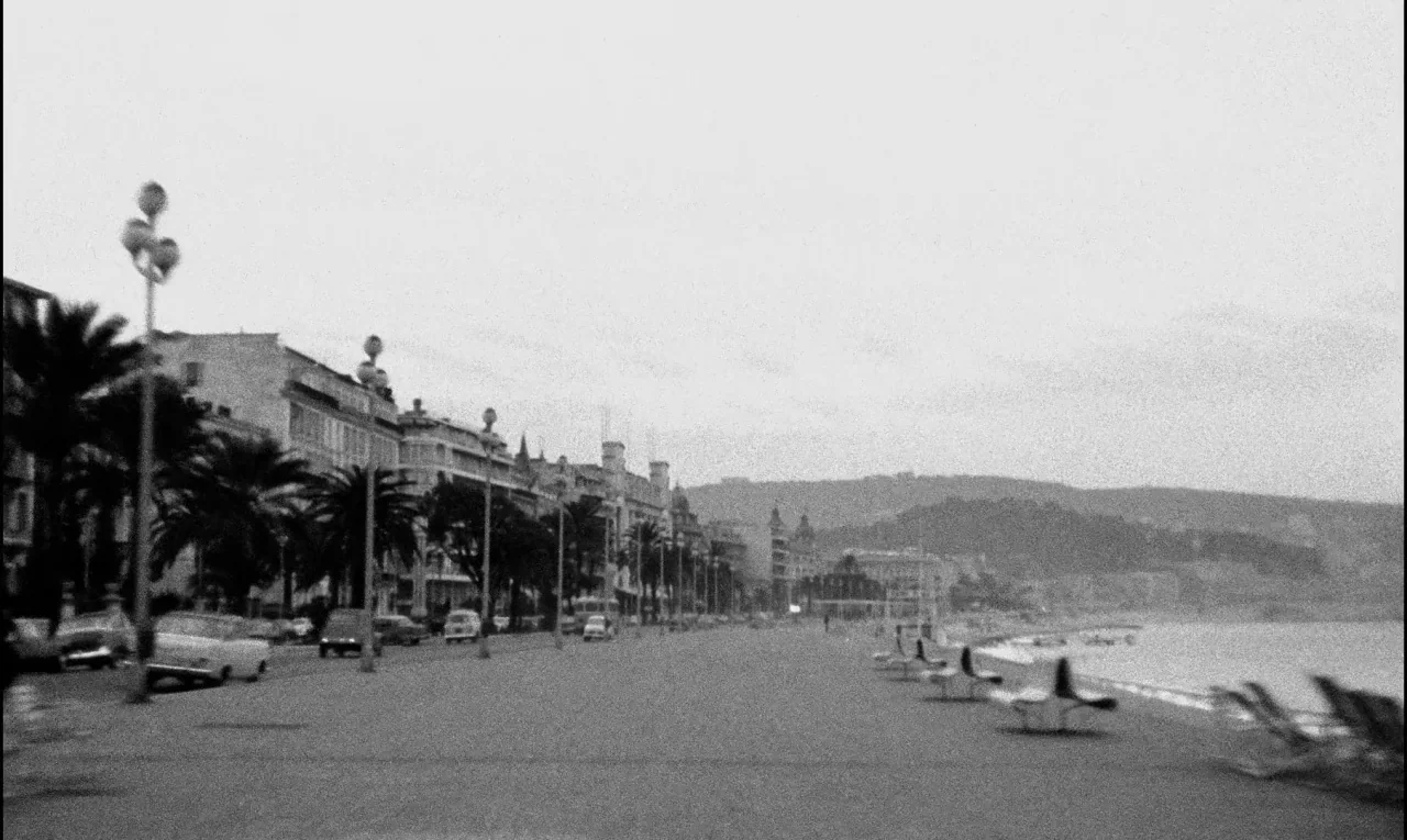 Bay of Angels (1963) movie still: wide — A long seaside promenade features classic street lamps and rows of parked cars. Empty benches line the path…