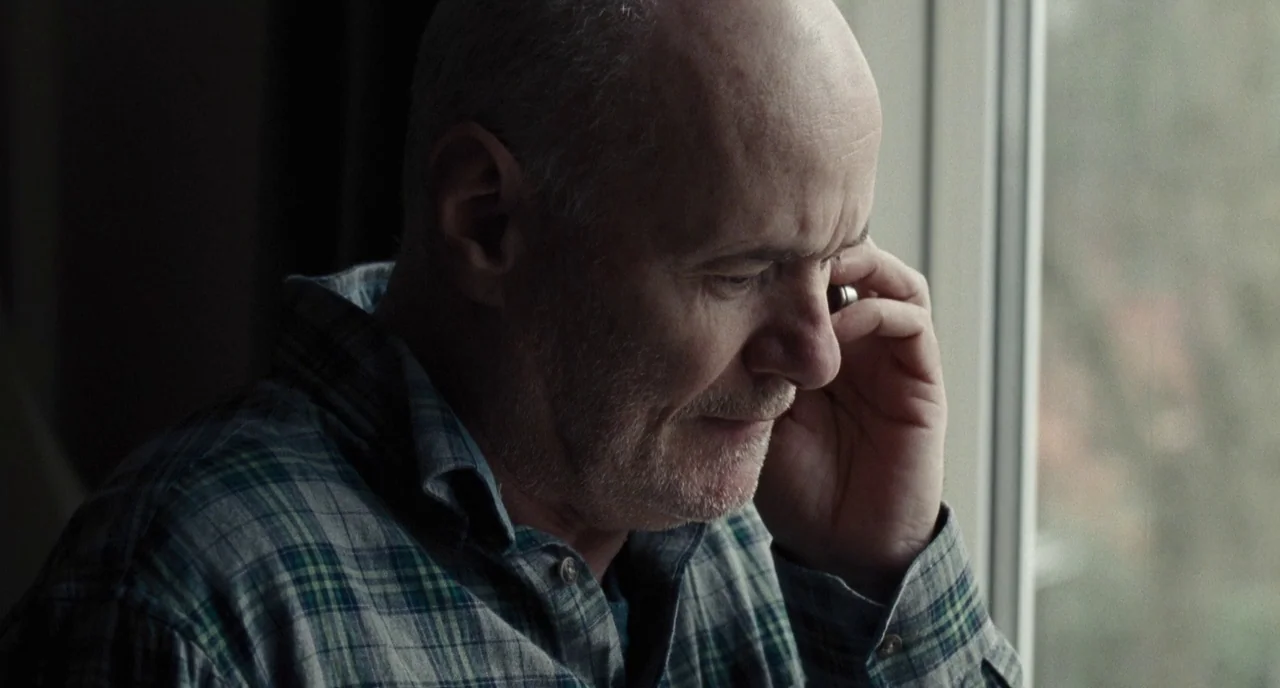 I, Daniel Blake (2016) movie still: closeup — A man stands indoors next to a large vertical window pane. He holds a small mobile phone to his ear…