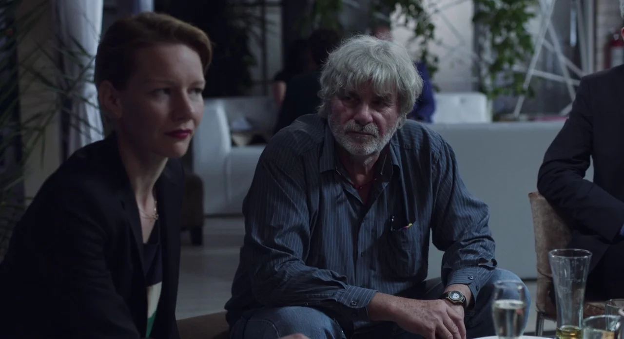 Toni Erdmann (2016) movie still: medium — A dimly lit lounge with soft seating and blurred interior architecture in the background. A woman in a…