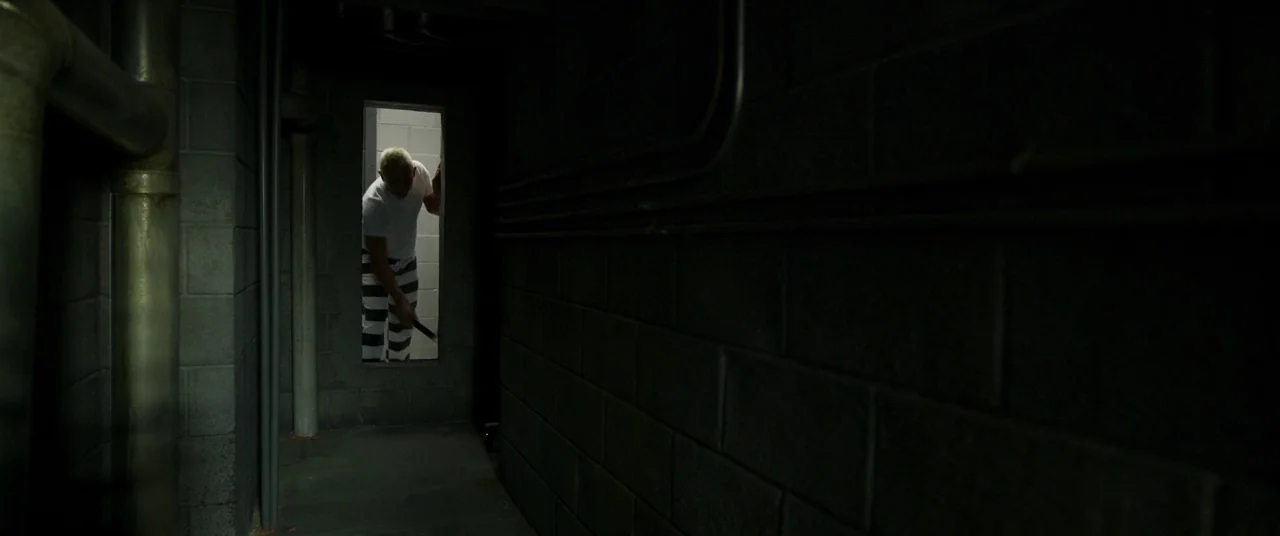 Logan Lucky (2017) movie still: wide — A dark hallway built from grey concrete blocks features a single illuminated doorway. A man wearing striped…