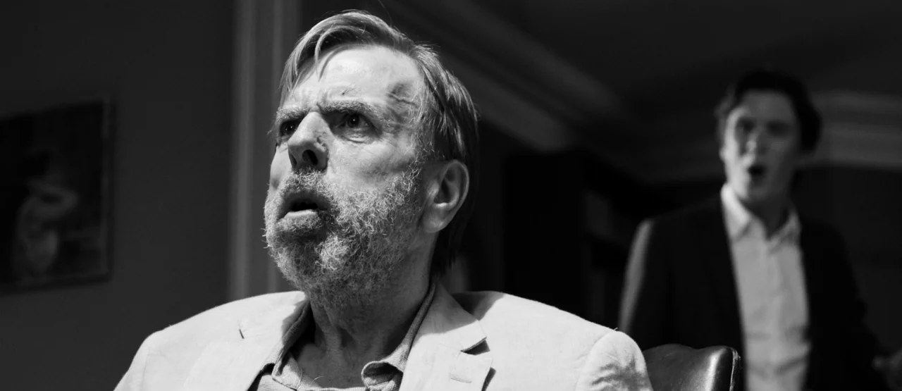 The Party (2017) movie still: medium — A dimly lit residential interior features an older man in the foreground and a younger man slightly out of…