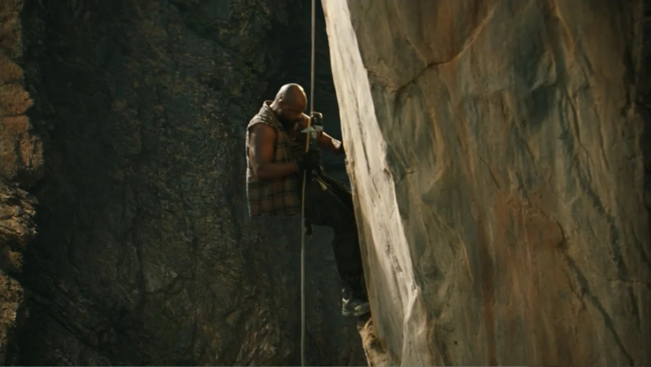 Jurassic World Rebirth (2025) movie still: fullbody — A man in a plaid vest descends a high, craggy rock face. He uses rappelling gear while…