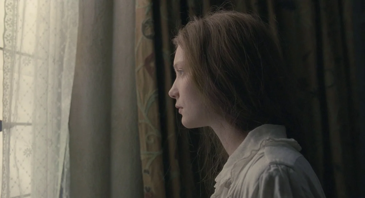 Jane Eyre (2011) movie still: closeup — A woman stands before a window draped with heavy patterned curtains and white lace. She gazes outward…