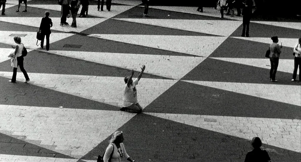 Container (2006) movie still: wide — A large public plaza features geometric white and dark gray triangular stone tiling patterns. A single person…