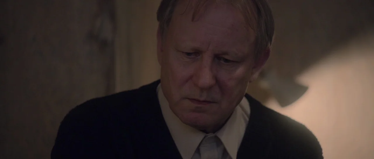 Nymphomaniac Vol 2 (2013) movie still: closeup — An interior space is dimly lit with beige walls and a soft light source. An older man with light…