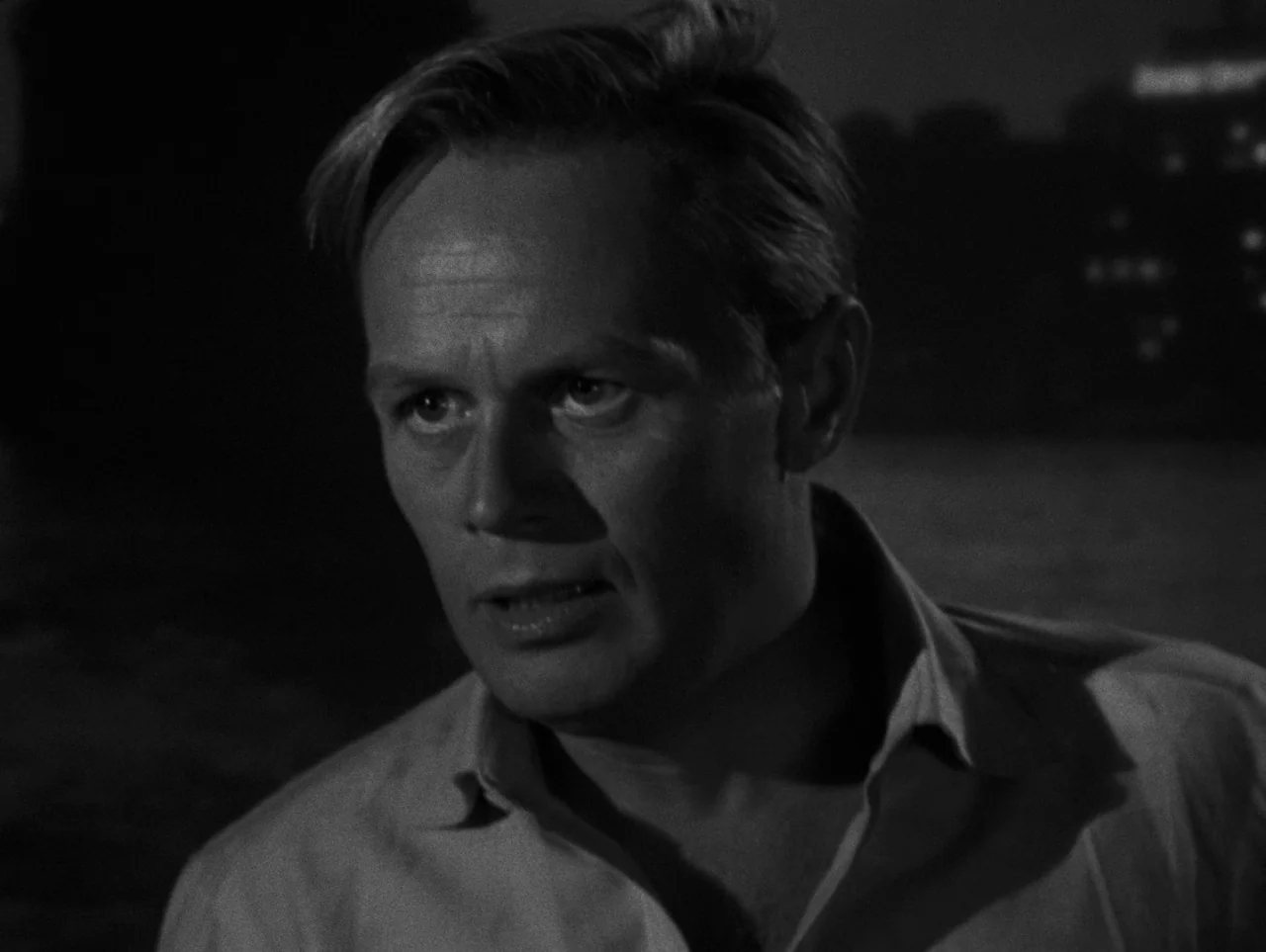 Pickup on South Street (1953) movie still: closeup — A dark outdoor space beside water with blurred city lights visible in the distance. A man turns…