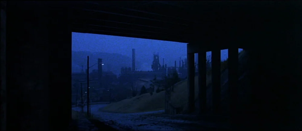 The Deer Hunter (1978) movie still: establishing — A view through a dark concrete underpass looking out toward a distant industrial complex. Smoke…