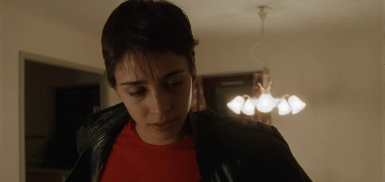 Pusher (1996) movie still: medium — A room with beige walls features a glowing multi-bulb chandelier suspended from the ceiling. A young woman with…