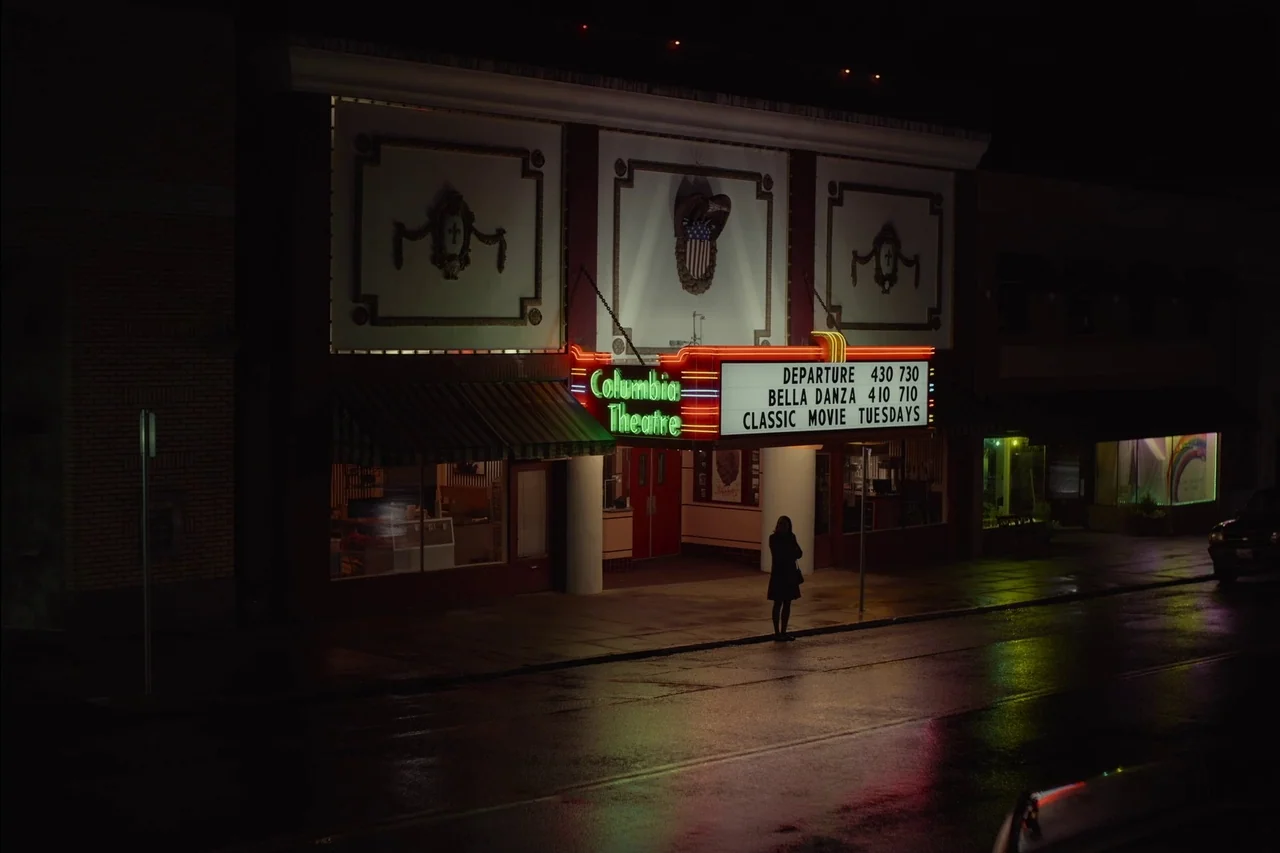 Sometimes I Think About Dying (2023) movie still: wide — An old-fashioned cinema facade with a neon sign glows brightly against a dark street. A…
