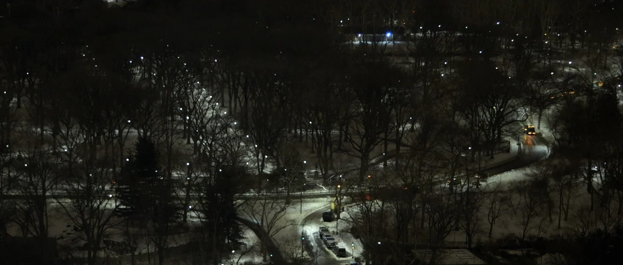 NY84 (2016) movie still: establishing — A snow-covered public park at night features winding roads and rows of bare trees. Scattered streetlights…