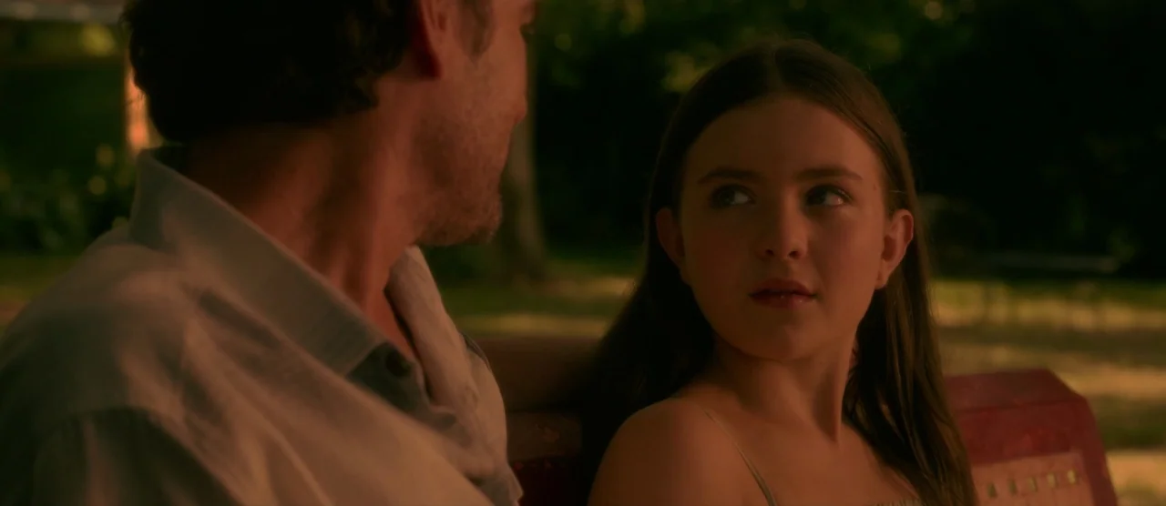 Gerald’s Game (2017) movie still: medium — Two people sit together on a park bench surrounded by blurred greenery. The girl looks toward the man…