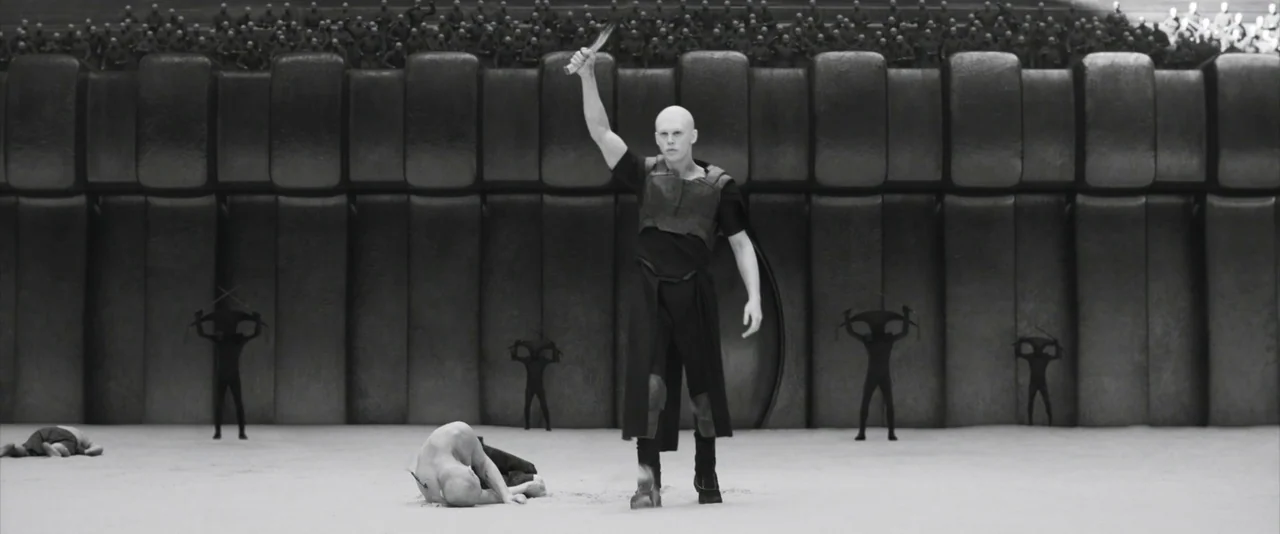 Dune: Part 2 (2024) movie still: fullbody — A vast, minimalist arena with tiered seating filled with a large, distant crowd. A bald man stands over…