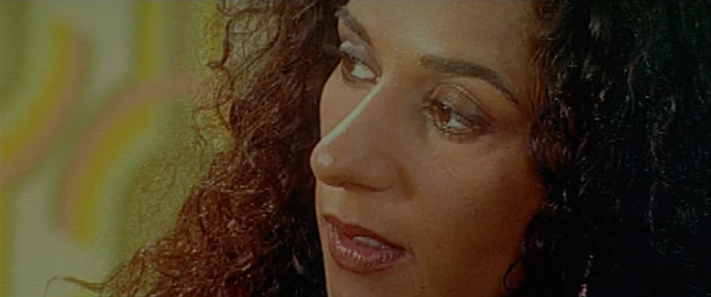 Talk To Her (2002) movie still: closeup — An interior space with a blurred, light-colored patterned background. A woman with dark curly hair looks…