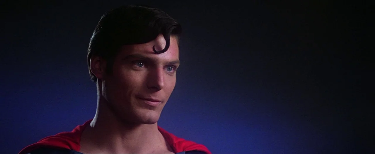 Superman (1978) movie still: closeup — A dark, indistinct space with a deep blue background gradient. A man in a blue suit and red cape turns his…