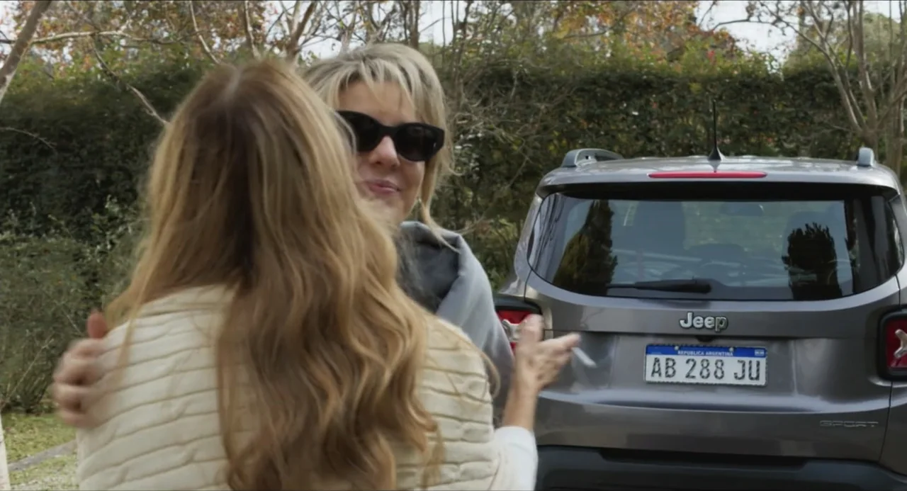 Echoes Of A Crime (2025) movie still: medium — A residential driveway with a grey Jeep parked in the background near trees. Two women stand together…